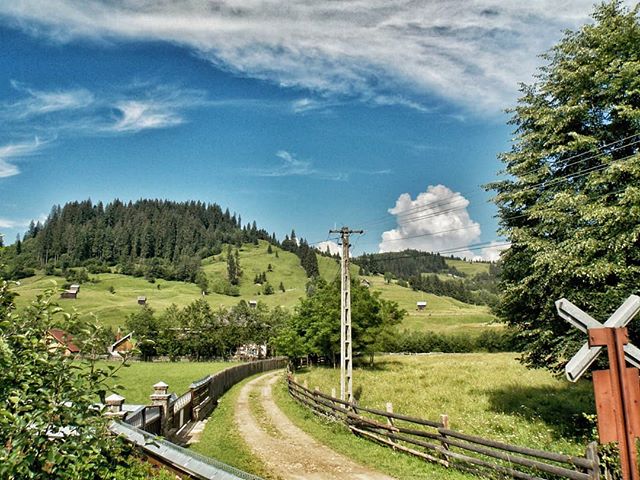 Scenic countryside view featuring rolling hills, a winding road, and lush greenery under a bright blue sky with clouds.