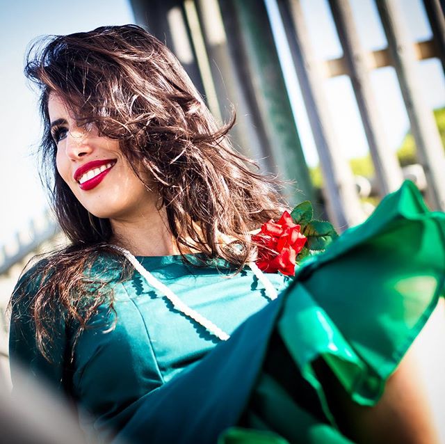 A smiling woman with red lipstick poses with a rose in a green dress, exuding confidence and charm.