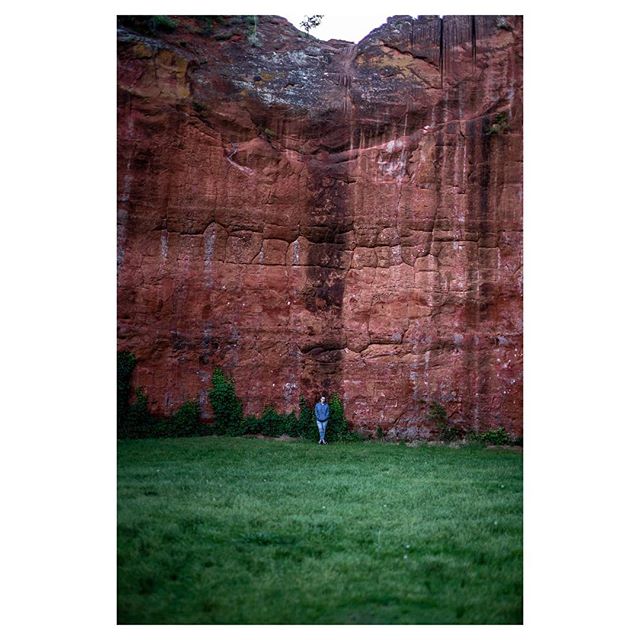 A lone figure stands before a massive red rock cliff surrounded by green grass, creating a sense of awe and tranquility.