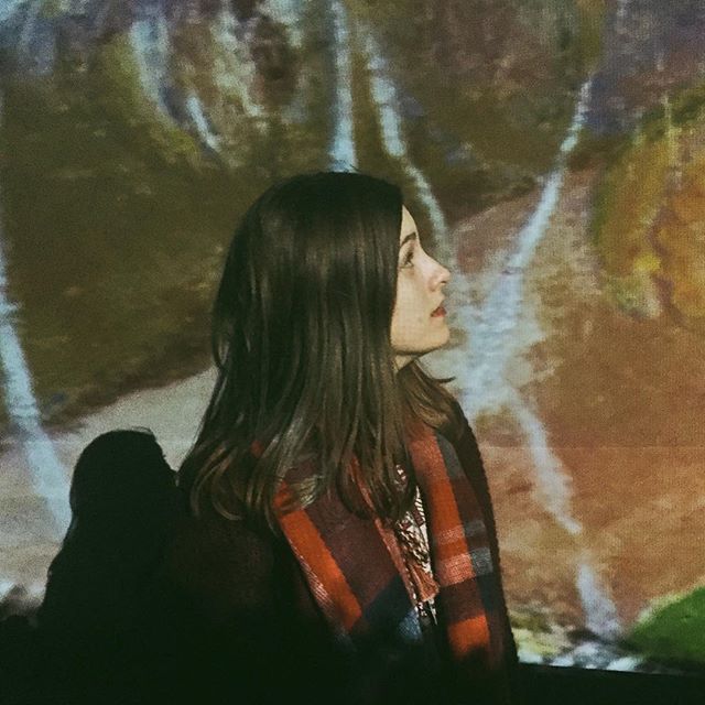 A woman wearing a scarf stands in front of an abstract art display, looking thoughtful.