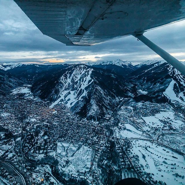 Aerial view of a snow-covered mountain town and ski resort under a cloudy sky from an airplane window.