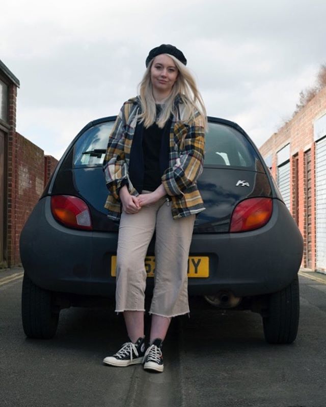 A blonde woman in a plaid shirt poses in front of a black Ford Ka in an urban alleyway.