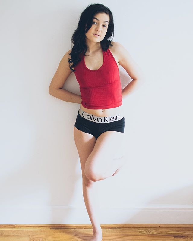 A young woman in a red tank top and Calvin Klein underwear poses against a white wall.