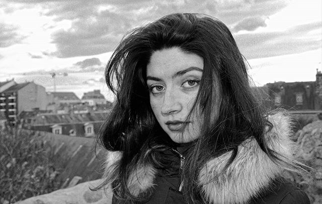 A young woman with dark hair poses in a black and white outdoor portrait with an urban backdrop.