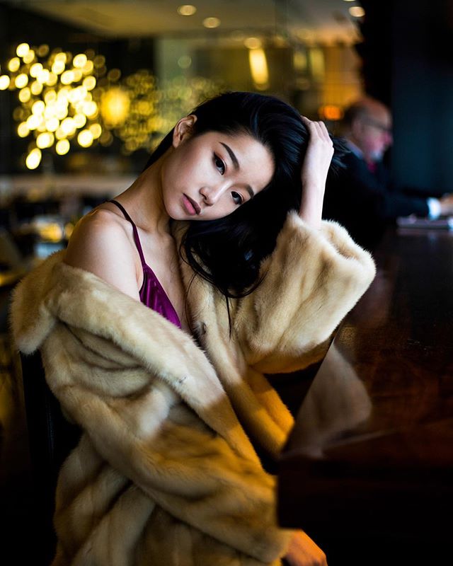 Elegant woman in fur coat poses in a bar, creating a sensual and dreamy editorial portrait.