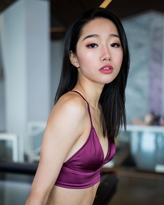 A young Asian woman poses in a purple top, looking sensual and calm in a soft, dreamy indoor setting.