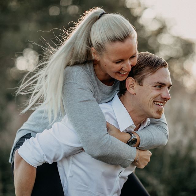 A happy couple enjoys a piggyback ride outdoors in a park, smiling and embracing the playful moment.