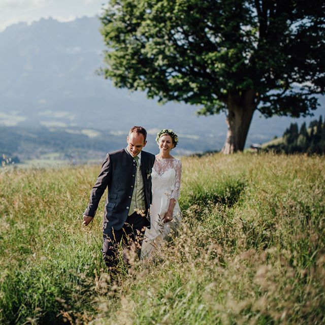 A bride and groom walk hand-in-hand through a grassy meadow with mountains in the background on their wedding day.
