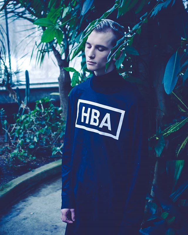 A man in a dark shirt with 'HBA' is surrounded by lush greenery in a botanical garden or greenhouse.