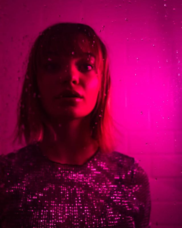 A woman with a sequined top stands against a vibrant pink background, creating a moody and mysterious portrait.