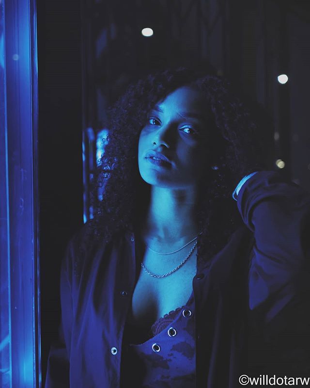 A woman with curly hair poses in a moody, blue-lit urban setting.