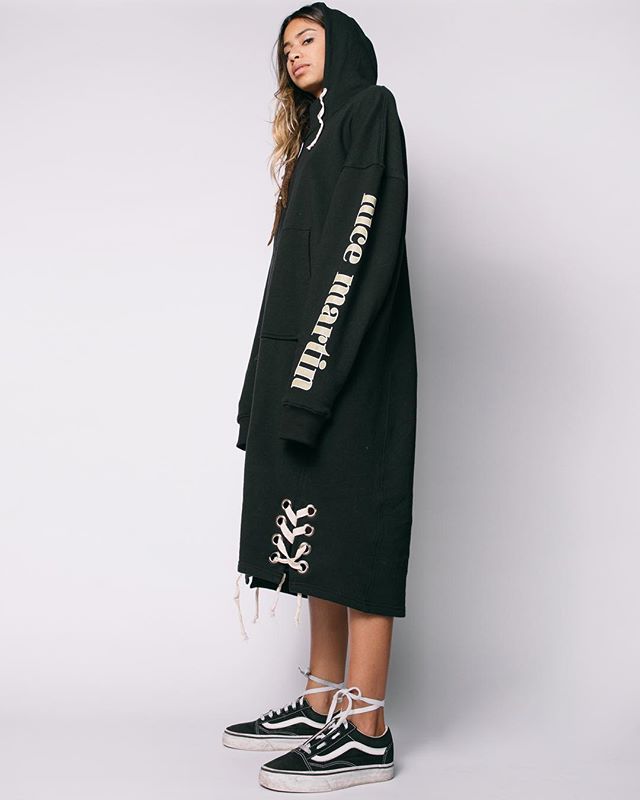 A woman models a black hoodie dress with white sneakers in a studio setting.