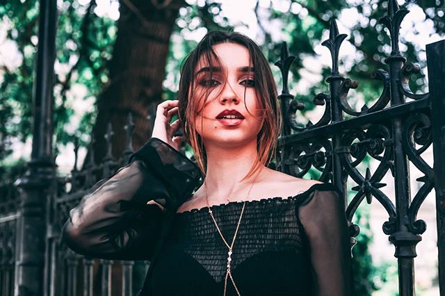 A young brunette woman poses in an off-shoulder top next to a wrought iron fence.
