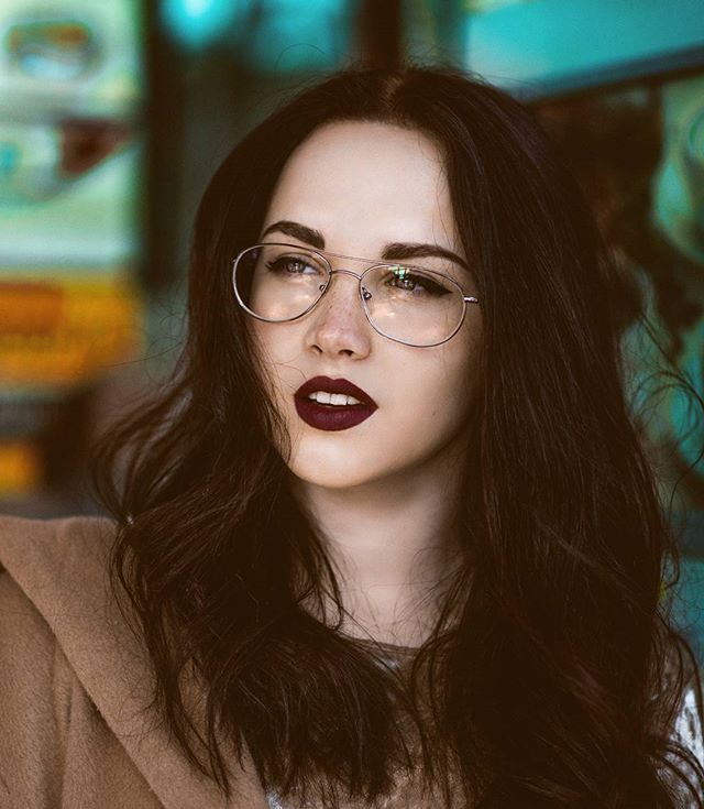 A brunette woman with glasses and dark lipstick poses in an urban setting.