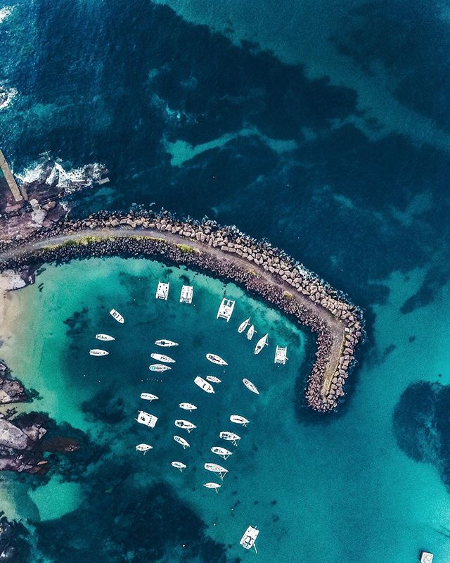 Aerial view of boats in a calm harbor with a rock breakwater along the coastline on a bright day.
