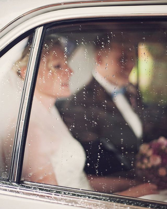 A bride and groom are seen through a rain-streaked car window on their wedding day.