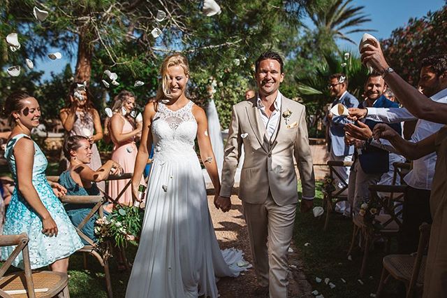 A bride and groom walk down the aisle amidst celebrating wedding guests throwing flower petals at an outdoor ceremony.