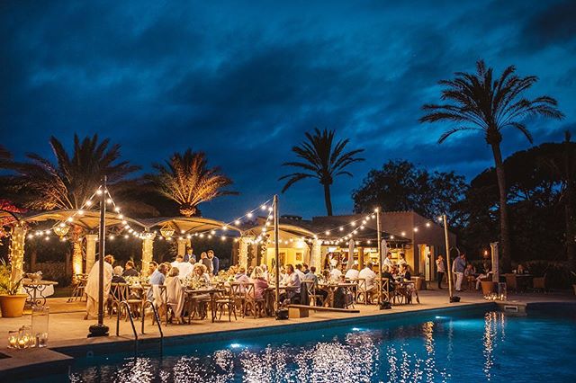 People are dining at an elegant outdoor party by the poolside under string lights at night.