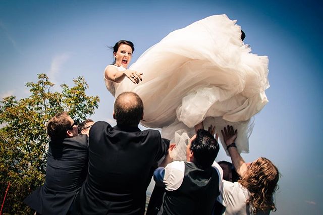 A joyous bride is lifted by the groomsmen, celebrating her wedding day under a bright blue sky.