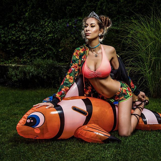 A woman poses on an inflatable fish wearing a bikini, shorts, and a floral jacket in a garden setting.