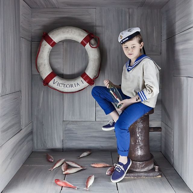 A boy dressed as a sailor sits with a message in a bottle surrounded by fish in a nautical themed studio.