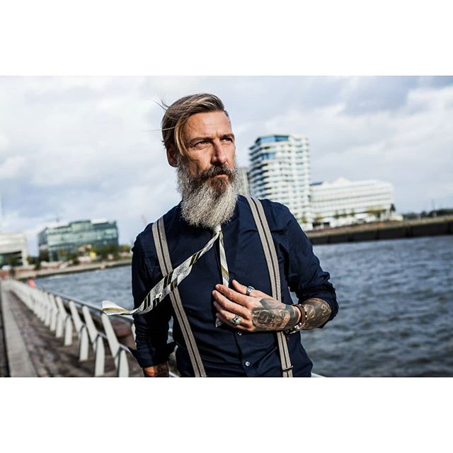 Stylish bearded man in suspenders and tie poses outdoors with waterfront city buildings in the background.