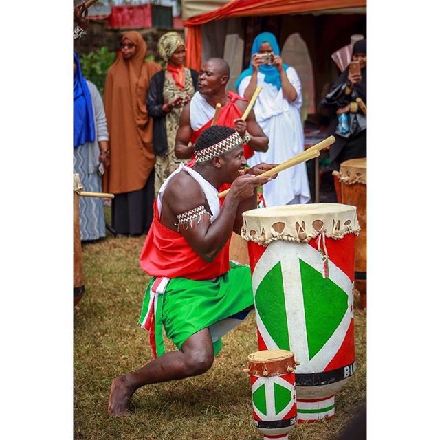 A drummer performs at a cultural event, wearing traditional clothing and playing drums with a lively, energetic style.
