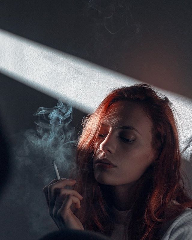 A red-haired woman is thoughtfully smoking a cigarette indoors, highlighted by soft light.