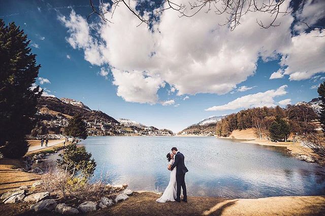 A bride and groom embrace by a serene lake surrounded by mountains on their wedding day.