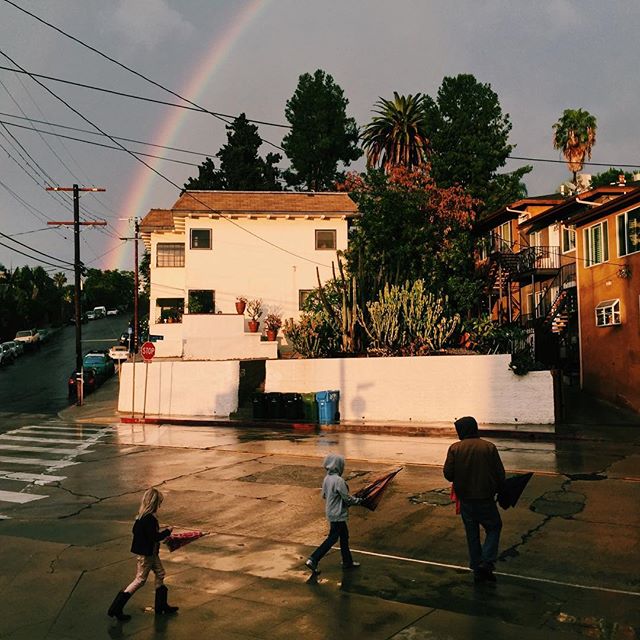 A father and two children walk across a wet street under a rainbow on a cloudy day.