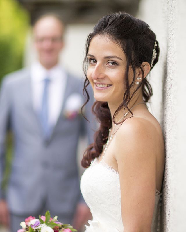 A smiling bride leans against a wall on her wedding day, with the groom blurred in the background.