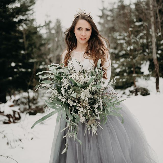A bride in a gray tulle dress holds a lush bouquet in a snowy winter forest.