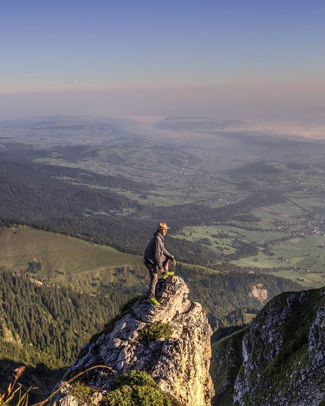 A man stands on a rocky mountain peak enjoying the view of the green valley below at sunrise.