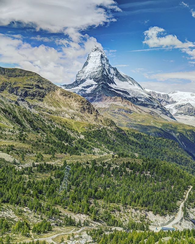 Scenic view of the Matterhorn mountain covered in snow, surrounded by green forests and a blue sky.