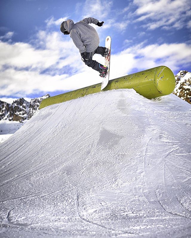 A snowboarder performs a trick on a rail in a snow park, set against a scenic mountain backdrop.