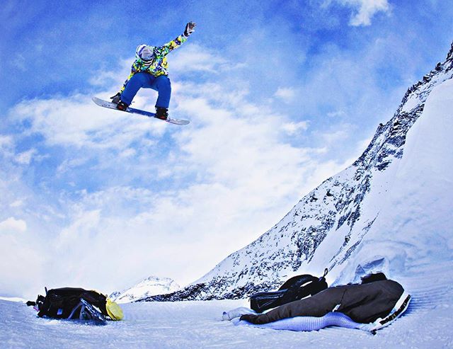 A snowboarder catches air against a scenic mountain backdrop, while another person relaxes in the snow.