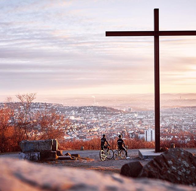Two cyclists pause by a large cross overlooking a European city at sunrise. 