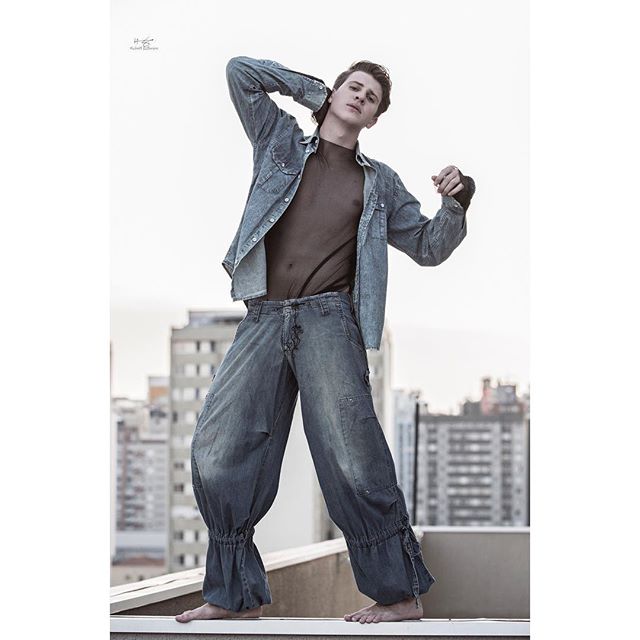 A stylish man poses on a rooftop in denim shirt and balloon pants against a city backdrop.