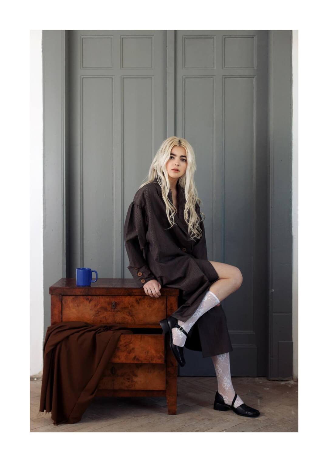 A woman with blonde hair poses in a brown dress, socks, and shoes, seated atop a wooden dresser.