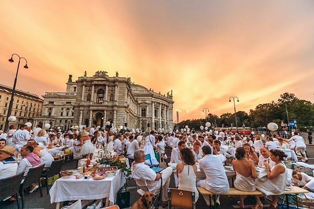 A large crowd gathers for a Dîner en Blanc-style dinner party in a city square at sunset.