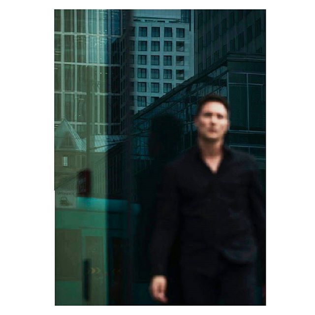 A man in black walks against a backdrop of modern city buildings. He looks serious, for potential business or fashion campaigns.