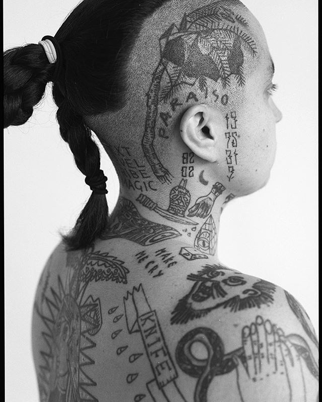 A person with many tattoos on their head, neck, and back, captured in a black and white studio portrait.