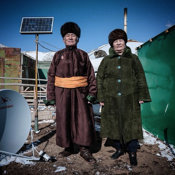 A traditionally dressed Mongolian couple poses outdoors near their home, showcasing their culture and lifestyle.