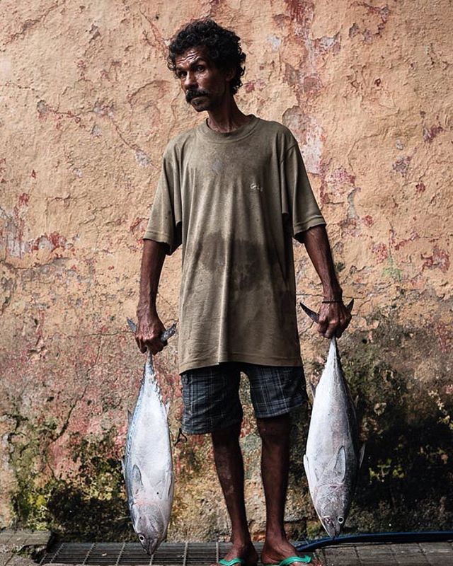 A fisherman stands with two fish against a textured wall.