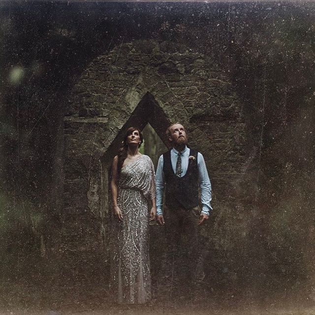 A formally dressed couple poses at a rustic ruin, looking upward, evoking a romantic and timeless feel.