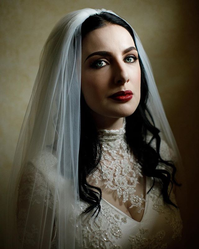 Elegant woman in wedding dress and veil with dark hair and red lipstick, looking at the camera.
