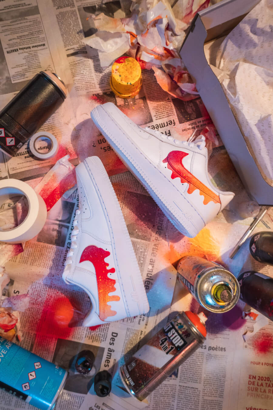 A pair of customized Nike Air Force 1 sneakers with red and orange spray paint accents, art supplies scattered around.