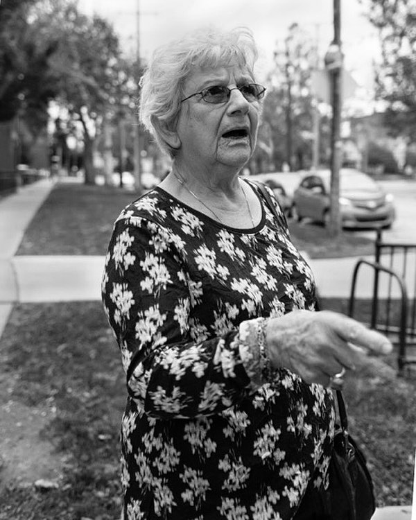 An elderly woman in a floral shirt stands outside, speaking and gesturing in a black and white portrait.