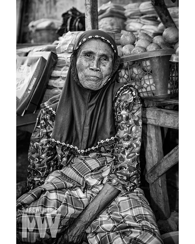 An elderly woman in a headscarf sits at a market, looking serene in a black and white portrait.