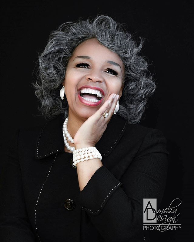 A cheerful woman with grey curly hair laughs against a black backdrop.